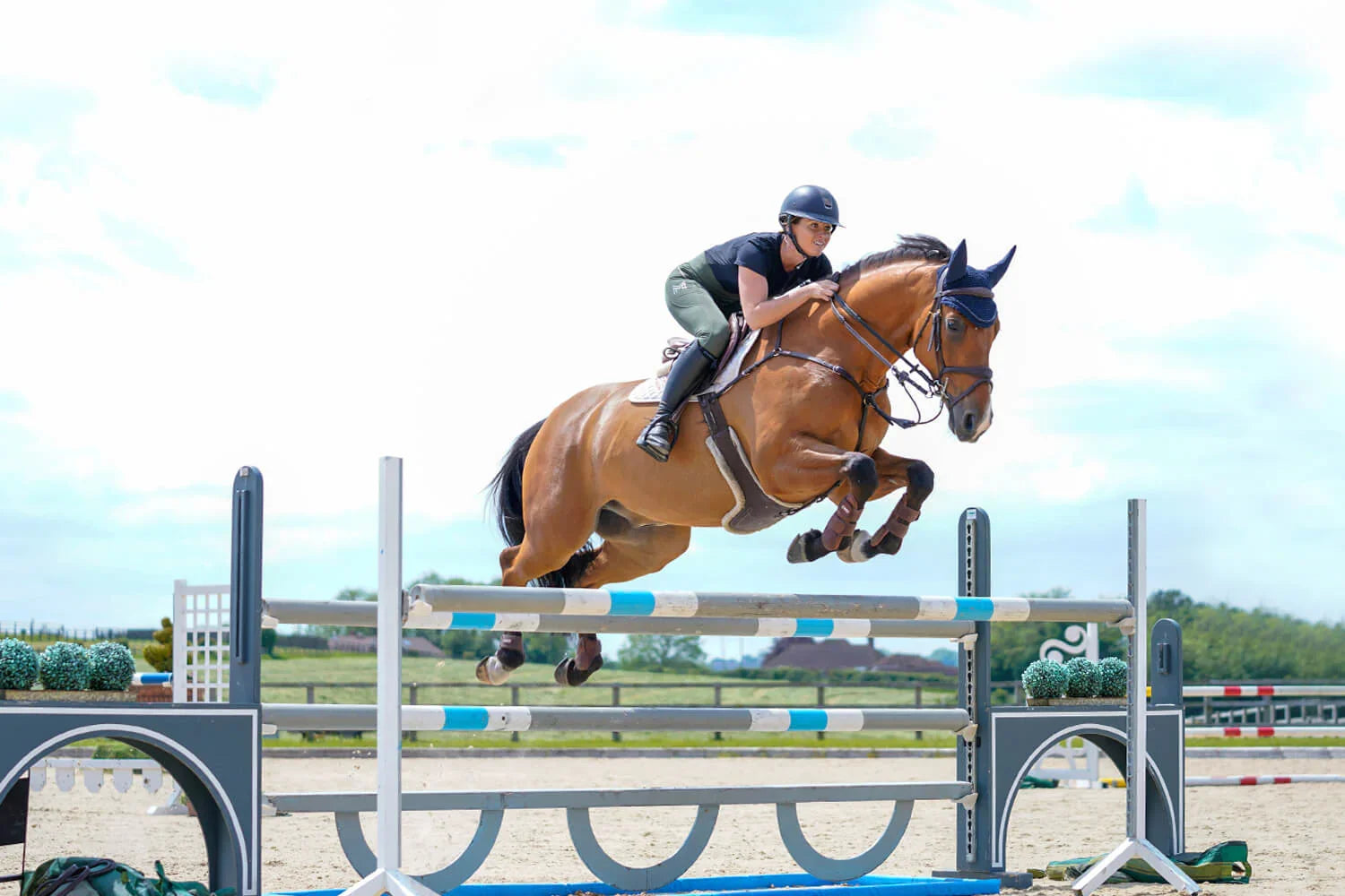 Reiterin springt mit braunem Pferd über ein Hindernis, Die #KAVALIOCANTERCHALLENGE – mit Stangenarbeit zum richtigen Rhythmus und Gleichmaß im Galopp.