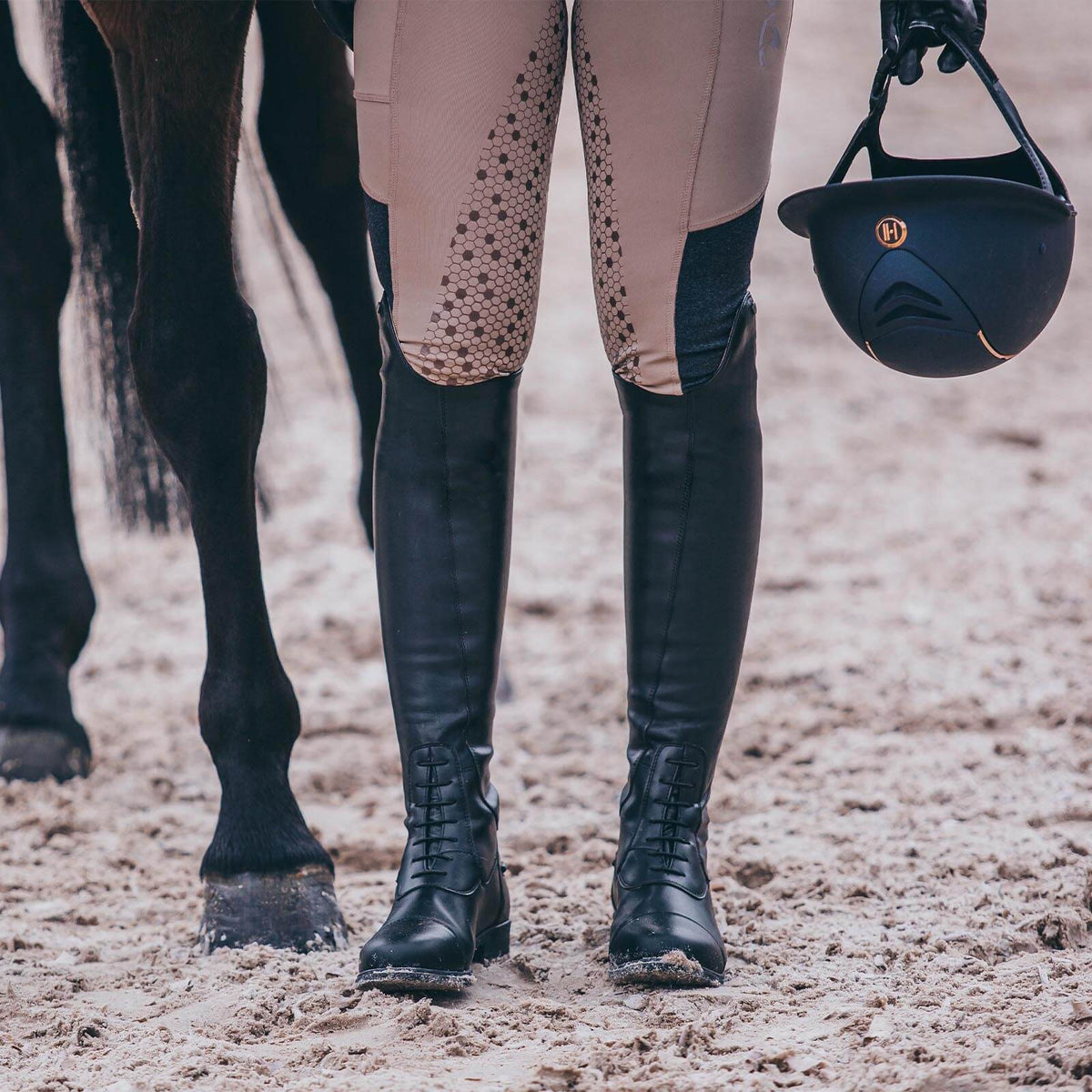 Suedwind Equestrian Reitstiefel Nova Tall Vegan, Schwarz, Person trägt sie neben einem Pferd im Reitplatz.